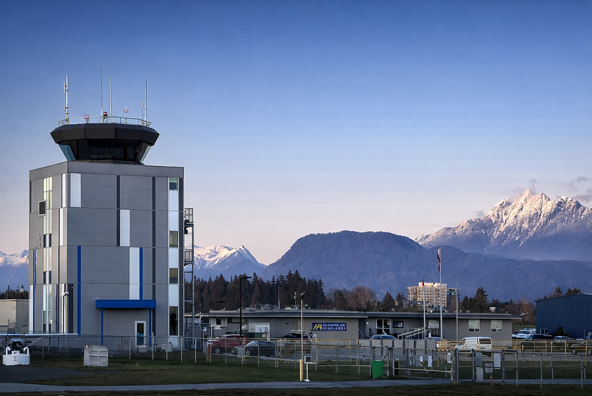 A NAV CANADA aviation control tower with a gray and blue exterior stands within a fenced operations site. Snow‑capped mountains and forested hills rise in the background under a clear sky.
